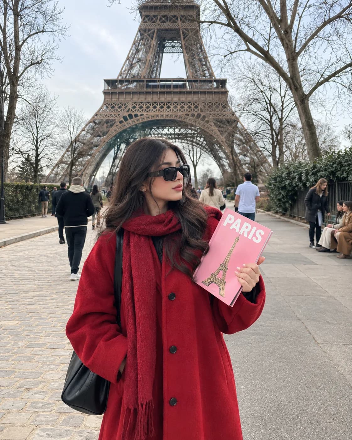A woman in a red coat holding a pink Paris photobook in front of the Eiffel Tower